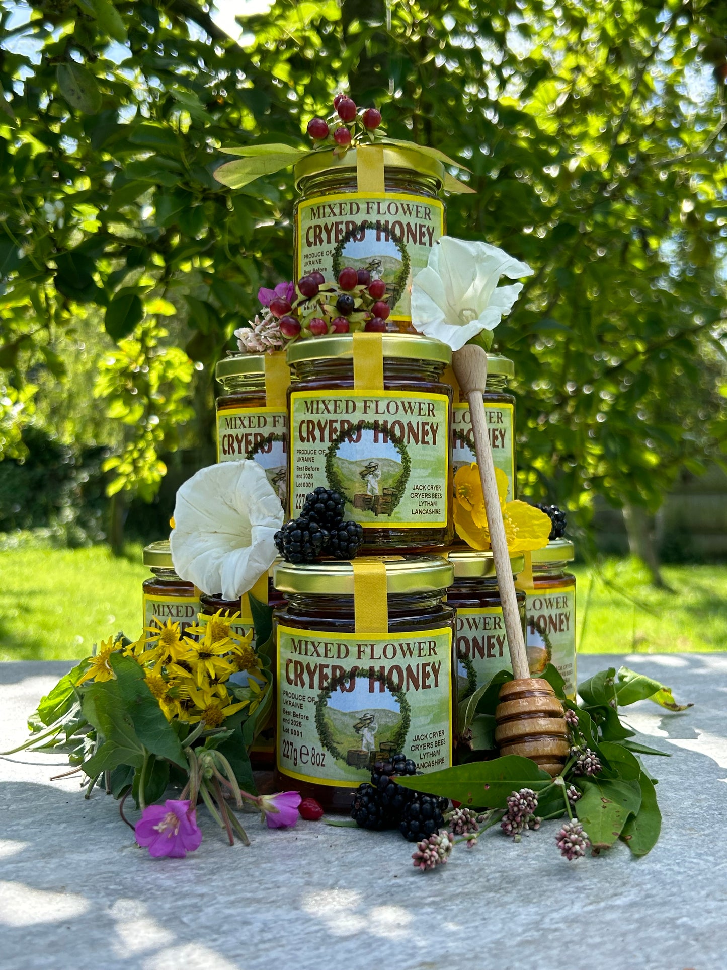 Stack of Ukrainian Mixed Flower Honey jars displayed outdoors with greenery in the background.