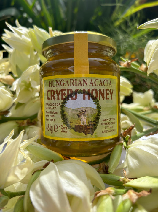 Close-up of a jar of Hungarian Acacia Honey by Cryer’s Honey, nestled among white flowers, showcasing its golden color and premium packaging.