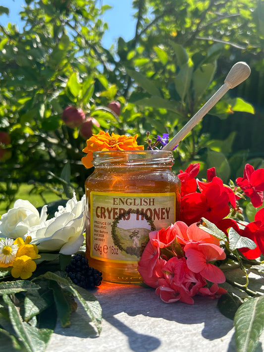 Jar of Cryer’s English Honey surrounded by vibrant flowers and greenery, with a honey dipper resting in the jar.