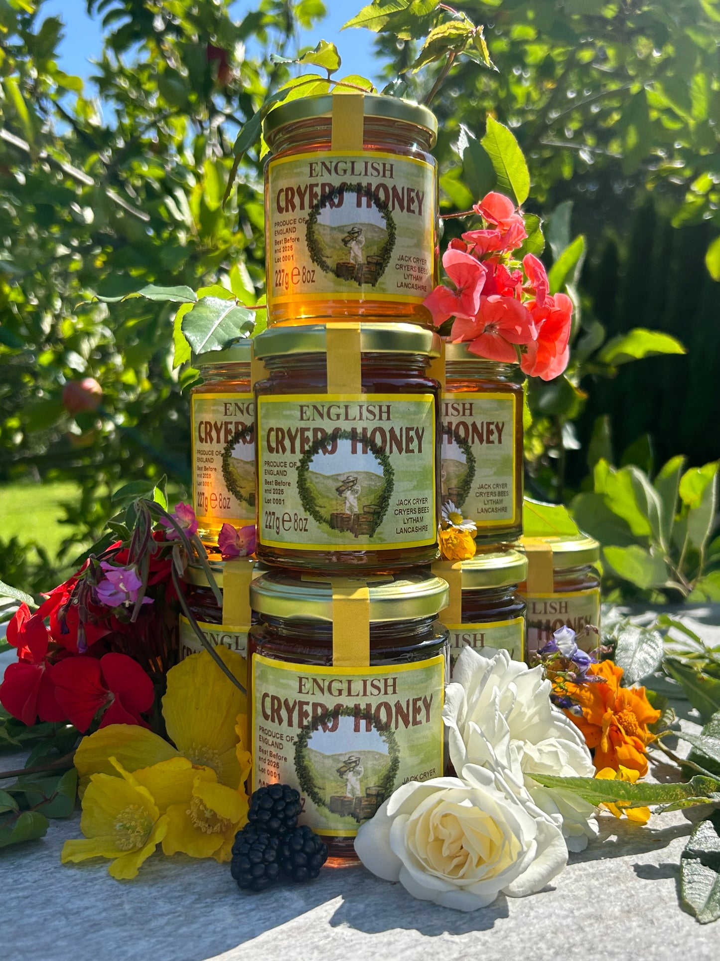 Pyramid of Cryer’s English Honey jars stacked in a garden, adorned with vibrant flowers and fresh blackberries.