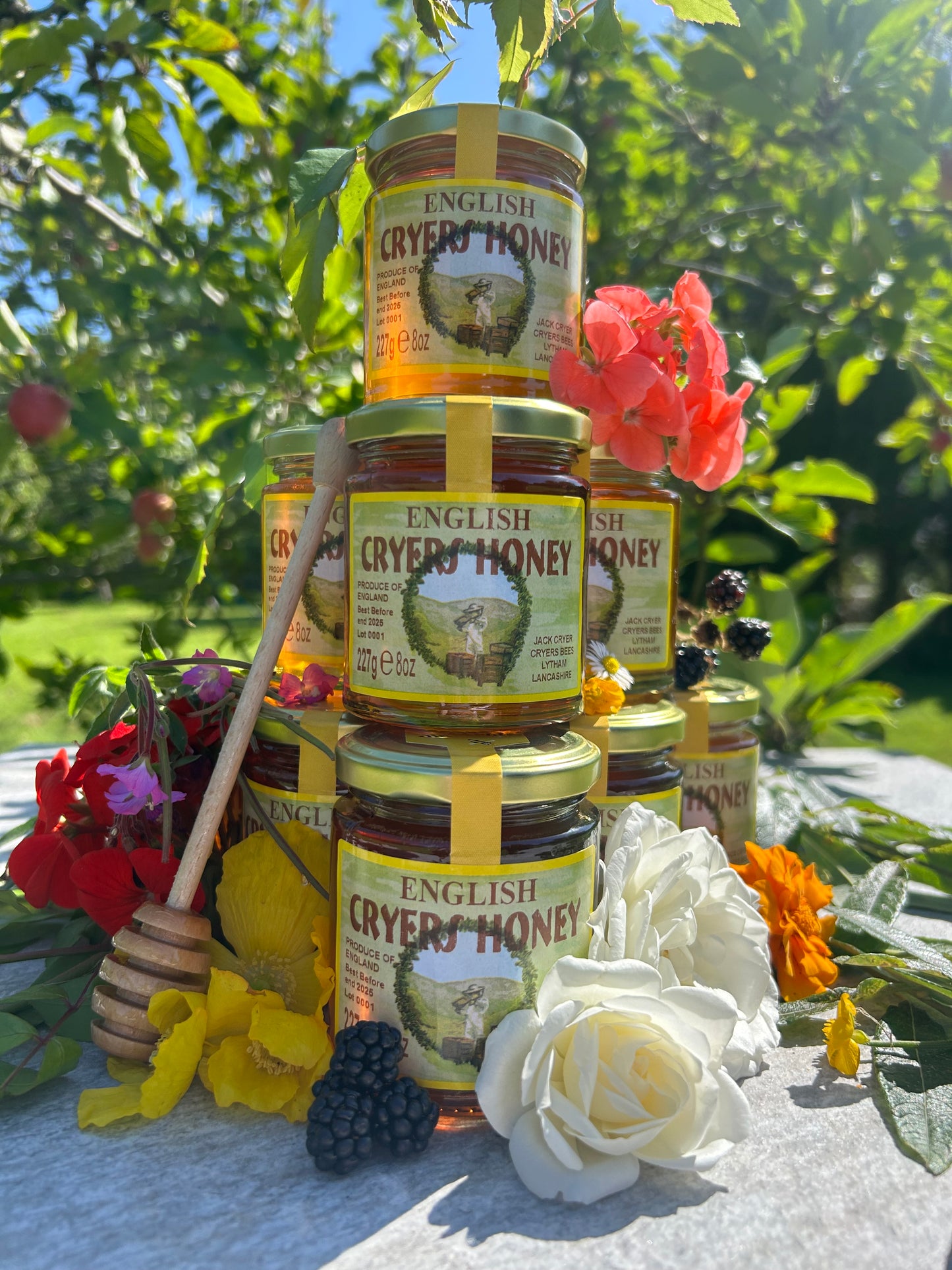 Stack of Cryer’s English Honey jars surrounded by flowers and blackberries, with a honey dipper resting against the jars in a lush garden setting.